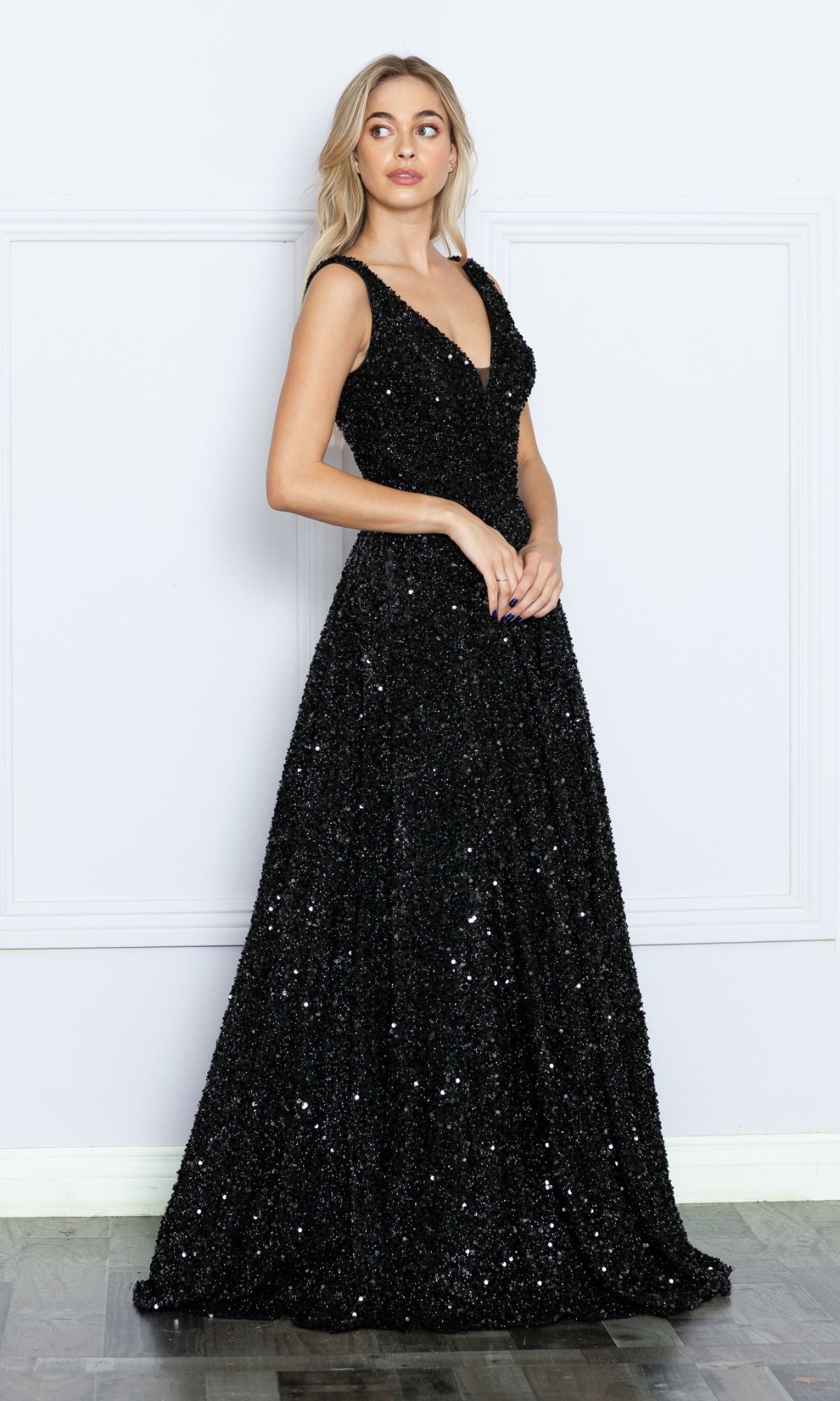 A woman stands indoors wearing the Long Black Sequin A-Line Prom Dress 9168, featuring a sleeveless design, V-neckline, fitted bodice, and floor-length sequin skirt.